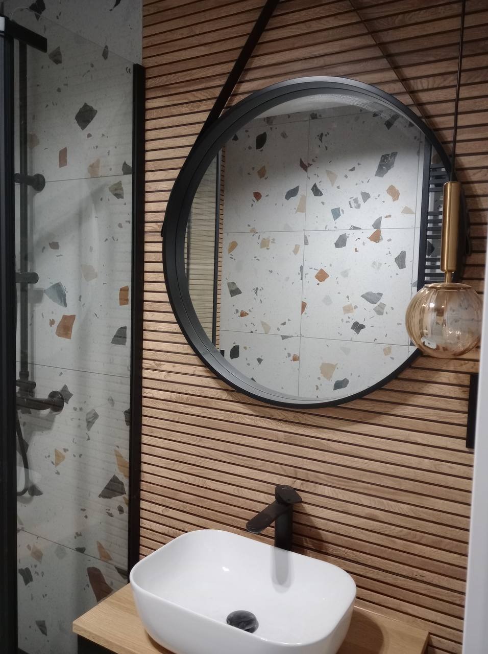 Nowoczesna łazienka z okrągłym lustrem, drewnianą boazerią i umywalką nablatową, widok na kabinę prysznicową z deszczownicą.
