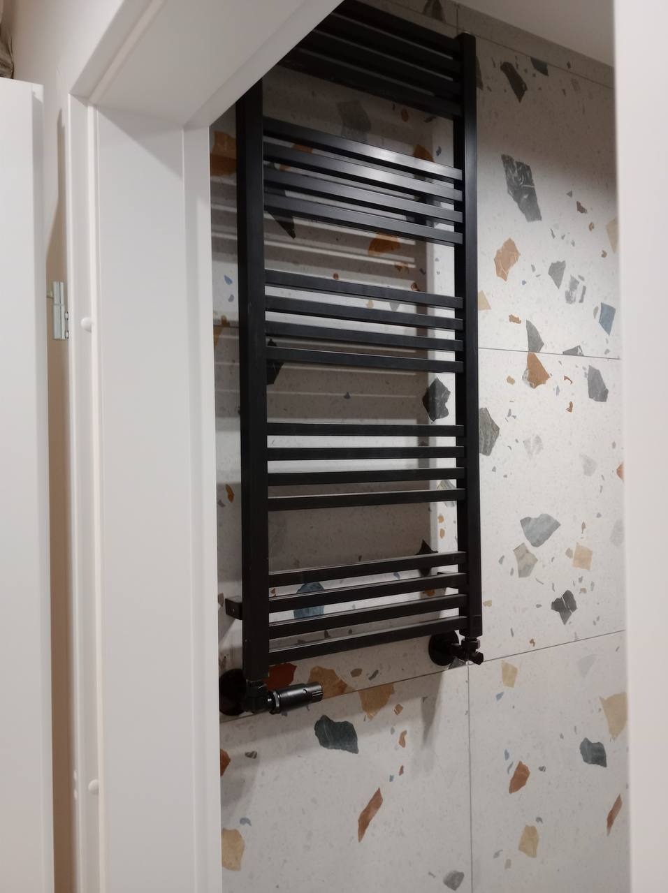 Czarny, drabinkowy grzejnik łazienkowy na ścianie z płytek imitujących lastryko, widoczny fragment białej ościeżnicy drzwi.