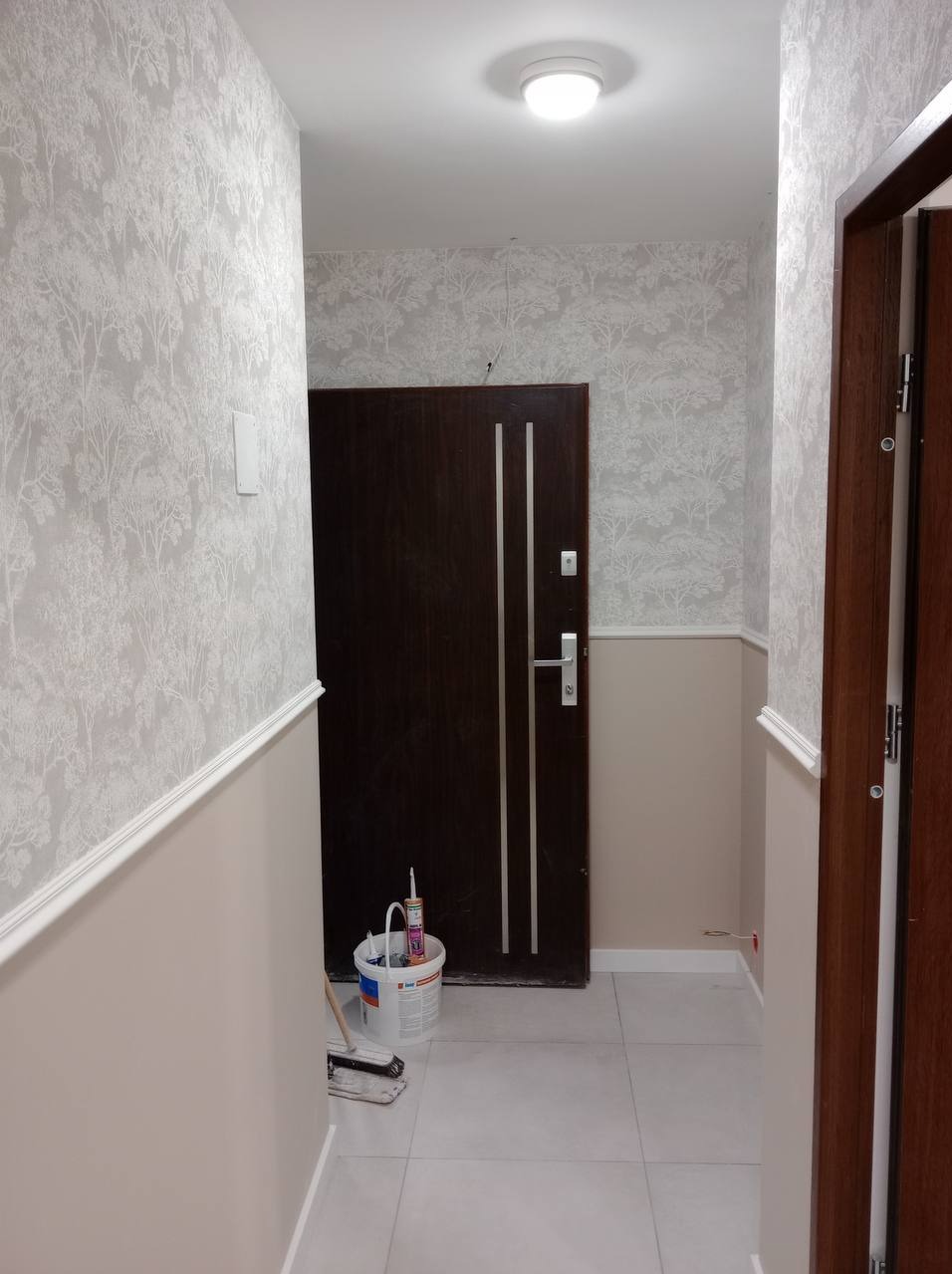 Korytarz po odświeżeniu ścian: dolna część pomalowana na beżowo, górna wytapetowana w delikatny wzór drzew, widoczne narzędzia malarskie przy drzwiach wejściowych.