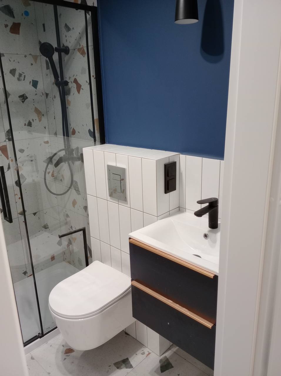 Nowoczesna łazienka z kabiną prysznicową o wzorzystych płytkach i podwieszaną toaletą, umywalka z szafką w kolorze ciemnego drewna z elementami w kolorze miedzi, ściana pomalowana na granatowo.