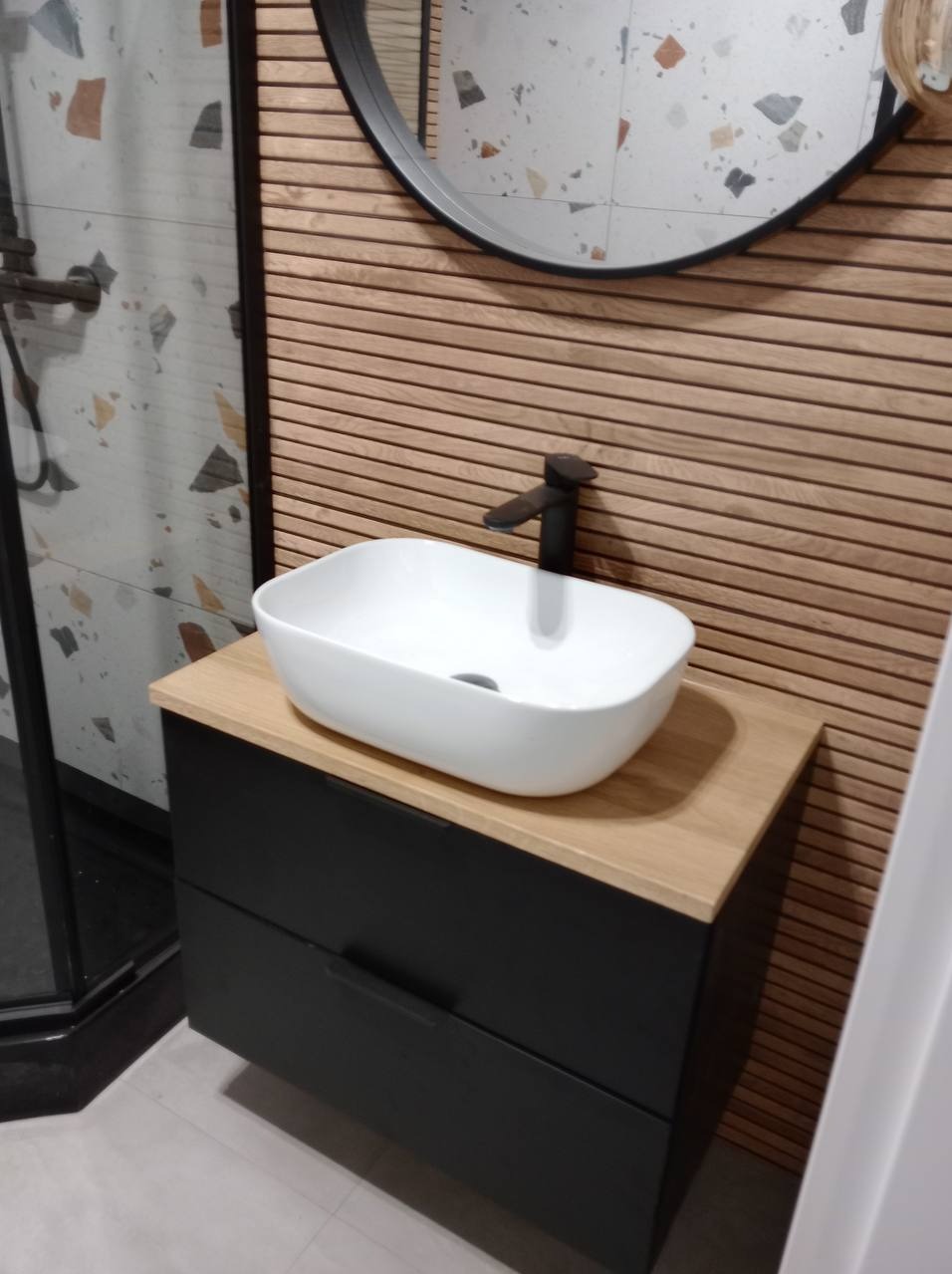 Nowoczesna łazienka z białą, owalną umywalką nablatową, czarną armaturą i szafką z szufladami w kolorze grafitowym, na tle ściany wyłożonej poziomymi panelami drewnianymi i lustra w czarnej ramie.
