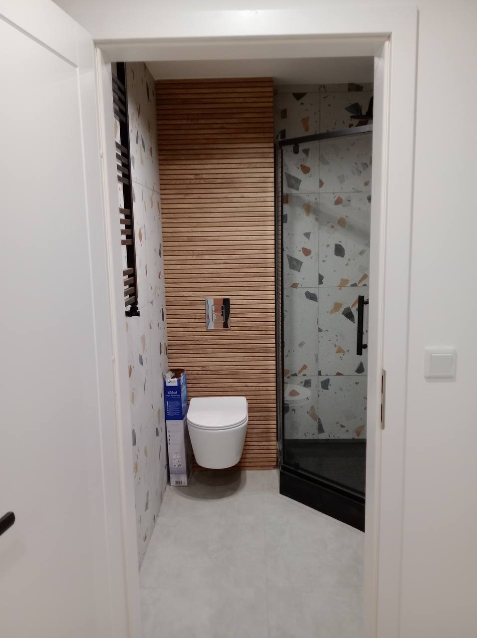 Nowoczesna łazienka z drewnianą ścianą z lameli, wiszącą toaletą i kabiną prysznicową z czarnymi ramami, widok zza otwartych drzwi.