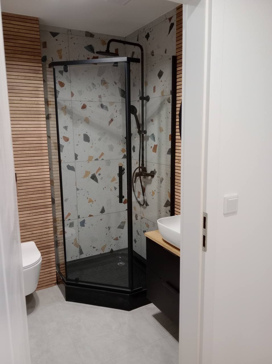 Nowoczesna łazienka z kabiną prysznicową w czarnej ramie, płytkami w stylu terrazzo, drewnianą ścianą i białą umywalką na czarnej szafce.