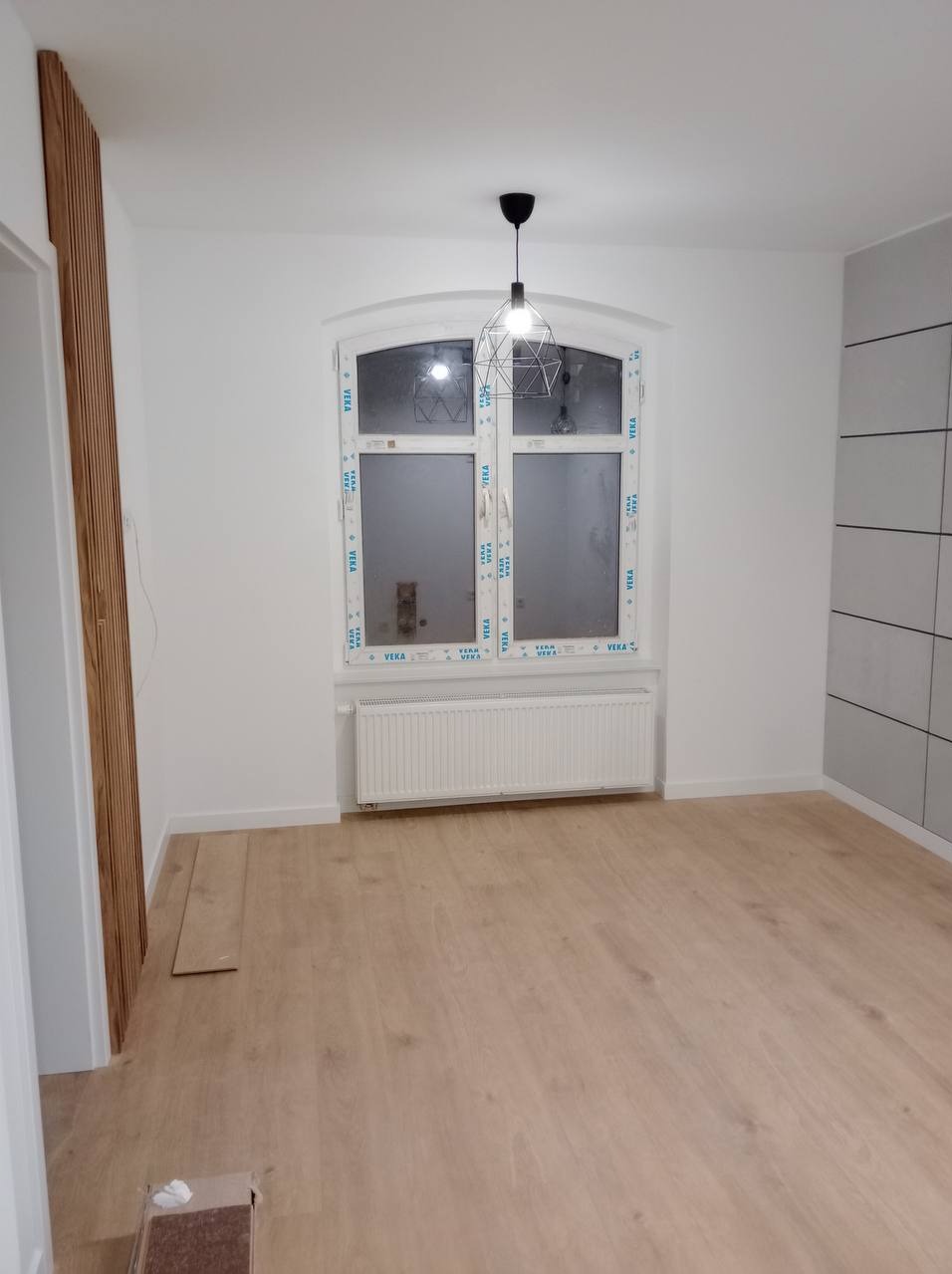 Wykończone wnętrze pokoju w Chorzowie po remoncie, z drewnianą podłogą, grzejnikiem pod łukowatym oknem i nowoczesną lampą wiszącą.