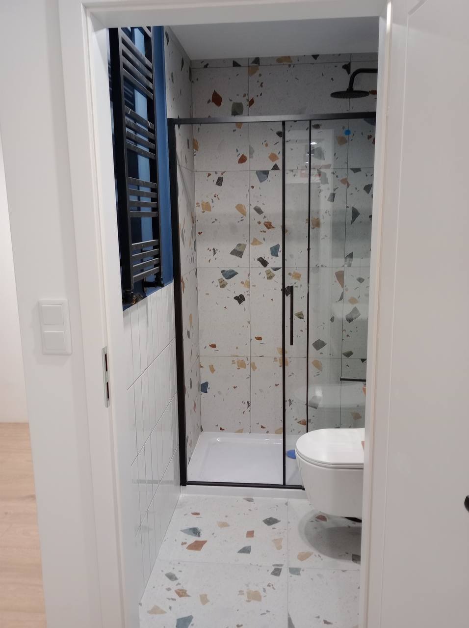 Wykończona łazienka z kabiną prysznicową z czarną ramą, białą armaturą i płytkami w stylu terrazzo na ścianach i podłodze; widok zza otwartych drzwi.