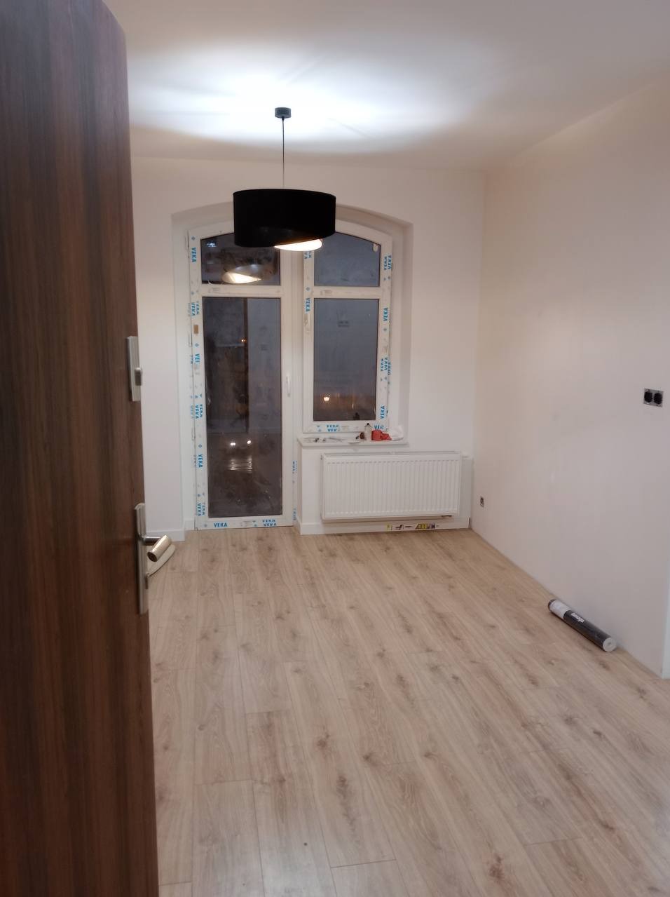 Pusty pokój po remoncie z nową podłogą panelową, świeżo pomalowanymi ścianami i zamontowanym grzejnikiem pod oknem. Na podłodze leży zwinięty rulon z folią. Drzwi wejściowe otwarte.