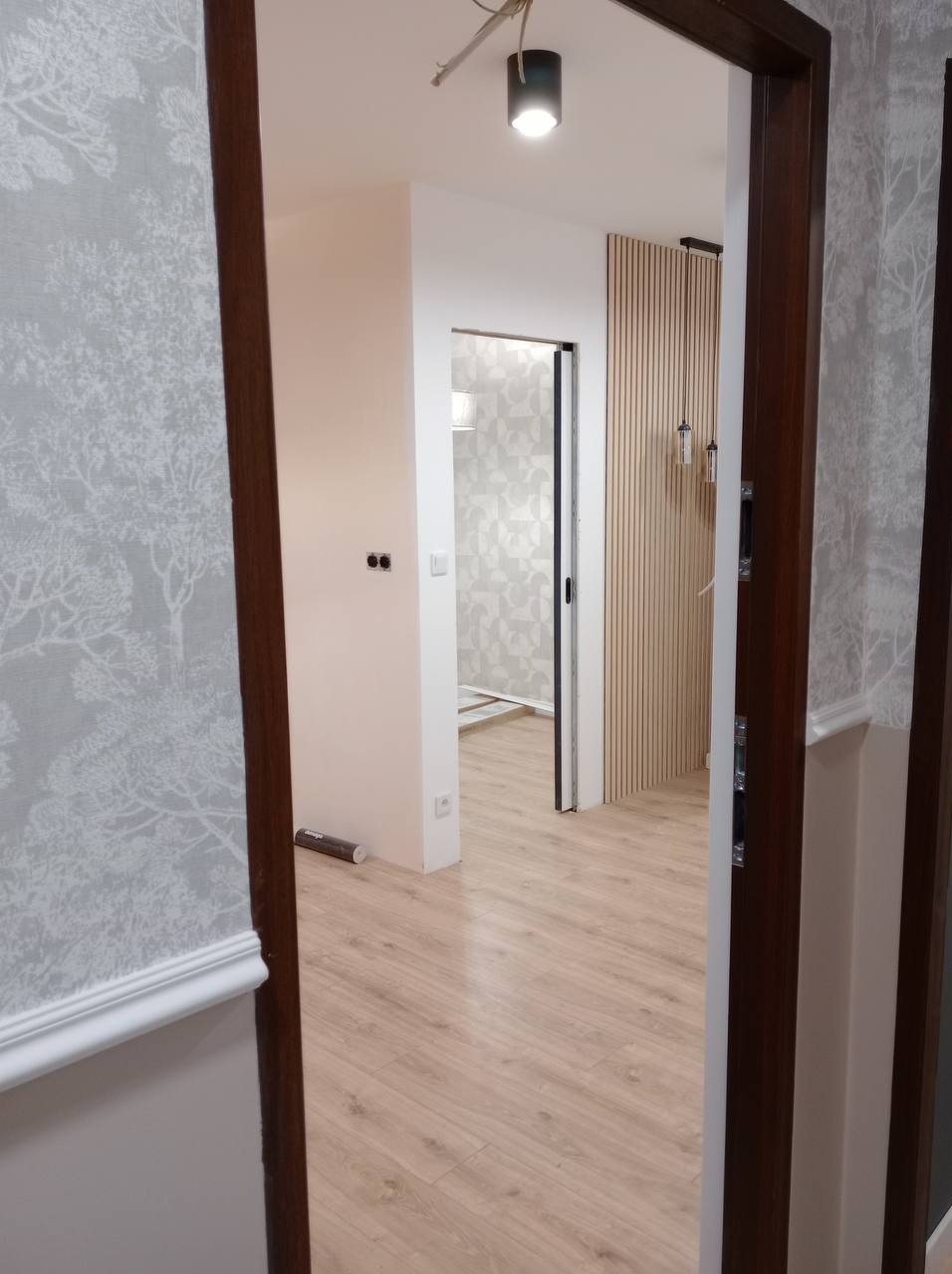 Widok na wykończone wnętrze mieszkania z nowymi panelami podłogowymi, tapetą w motywy drzew i ozdobną ścianą z drewnianych listew.