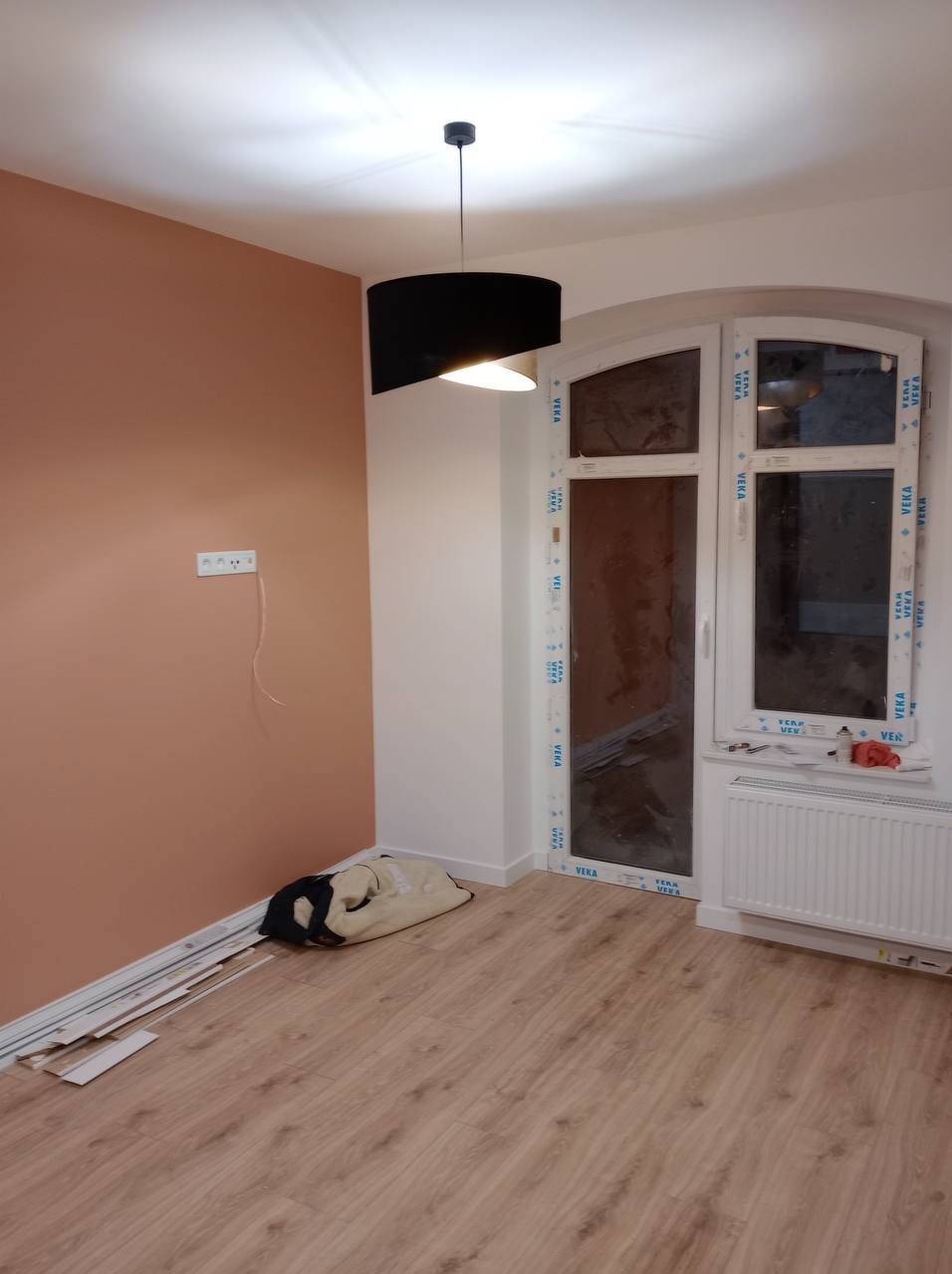 Pokój po odświeżeniu: ściana w odcieniach brzoskwini, nowe panele podłogowe, okno z białymi ramami, czarna lampa sufitowa. Widoczne listwy przypodłogowe i torba na podłodze.