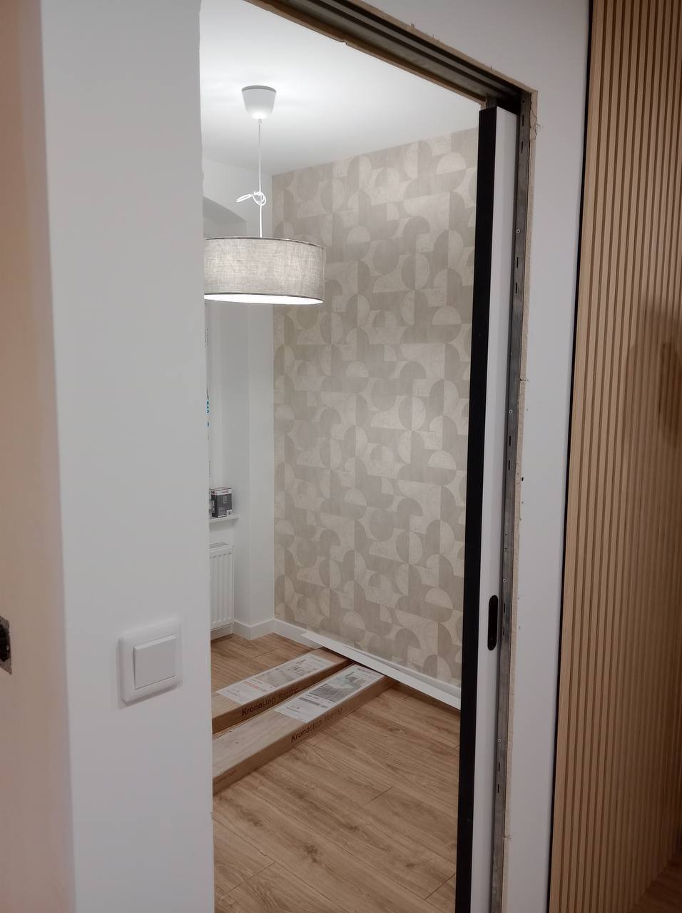 Wnętrze pokoju w trakcie wykańczania, widoczne panele podłogowe Kronostep w opakowaniach, ściana z wzorzystą tapetą i lampa sufitowa, obok ściana z drewnianymi lamelami. Montaż drzwi przesuwnych.