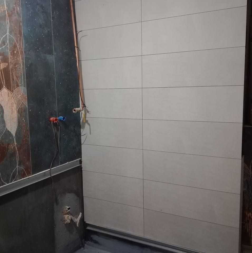 Ściana łazienki w trakcie wykańczania: ułożone jasne, prostokątne płytki ceramiczne na jednej ścianie, surowy tynk i instalacja wodna na drugiej.
