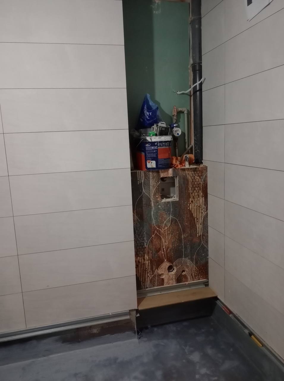 Remont łazienki w trakcie: częściowo wyłożone płytki ścienne, odsłonięty fragment instalacji, wiadro z farbą i narzędzia malarskie na ozdobnej obudowie.