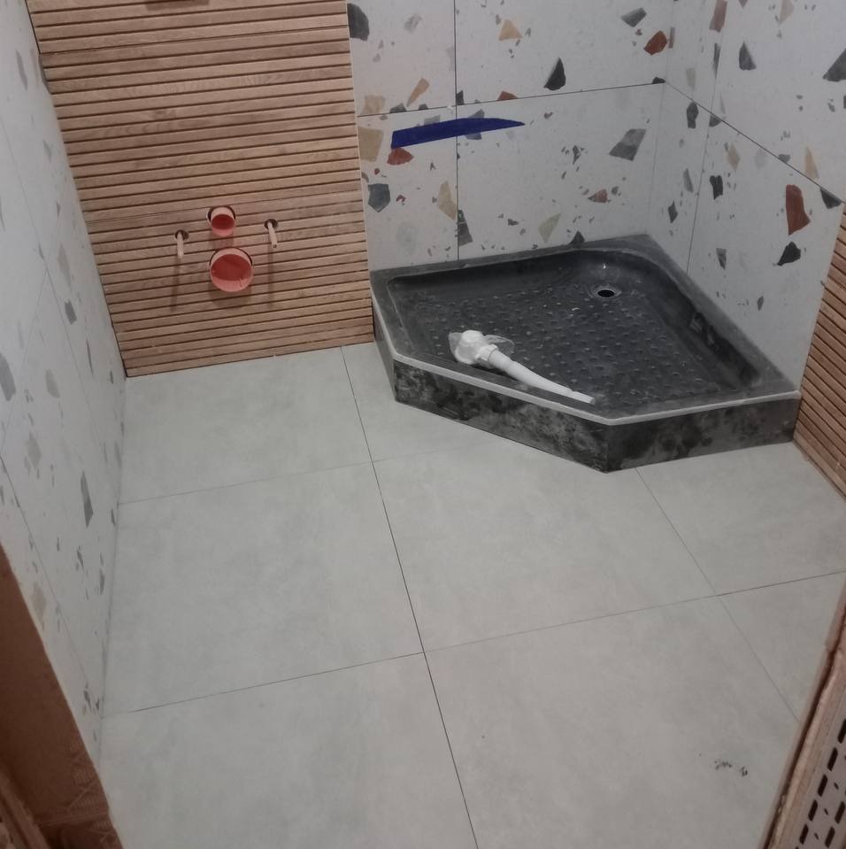 Narożny brodzik z szarego kamienia w trakcie montażu w łazience z płytkami imitującymi drewno i mozaikę, widoczny syfon.