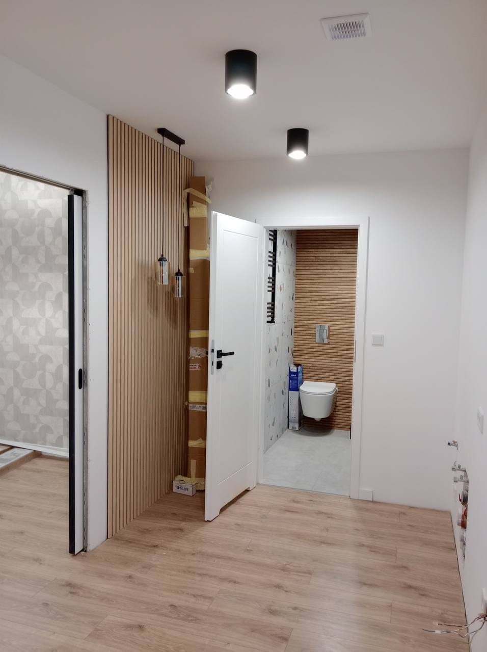 Wnętrze mieszkania w trakcie remontu: widok na wykończoną łazienkę z drewnianą okładziną, panele ścienne w przedpokoju i kartony z materiałami budowlanymi przy ścianie.