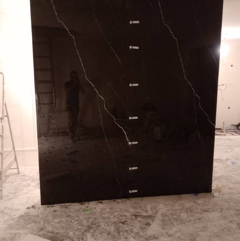 Czarna płyta z marmurkowym wzorem, zamocowana do ściany za pomocą tymczasowych kołków dystansowych, widoczna drabina i odbicie osoby robiącej zdjęcie.