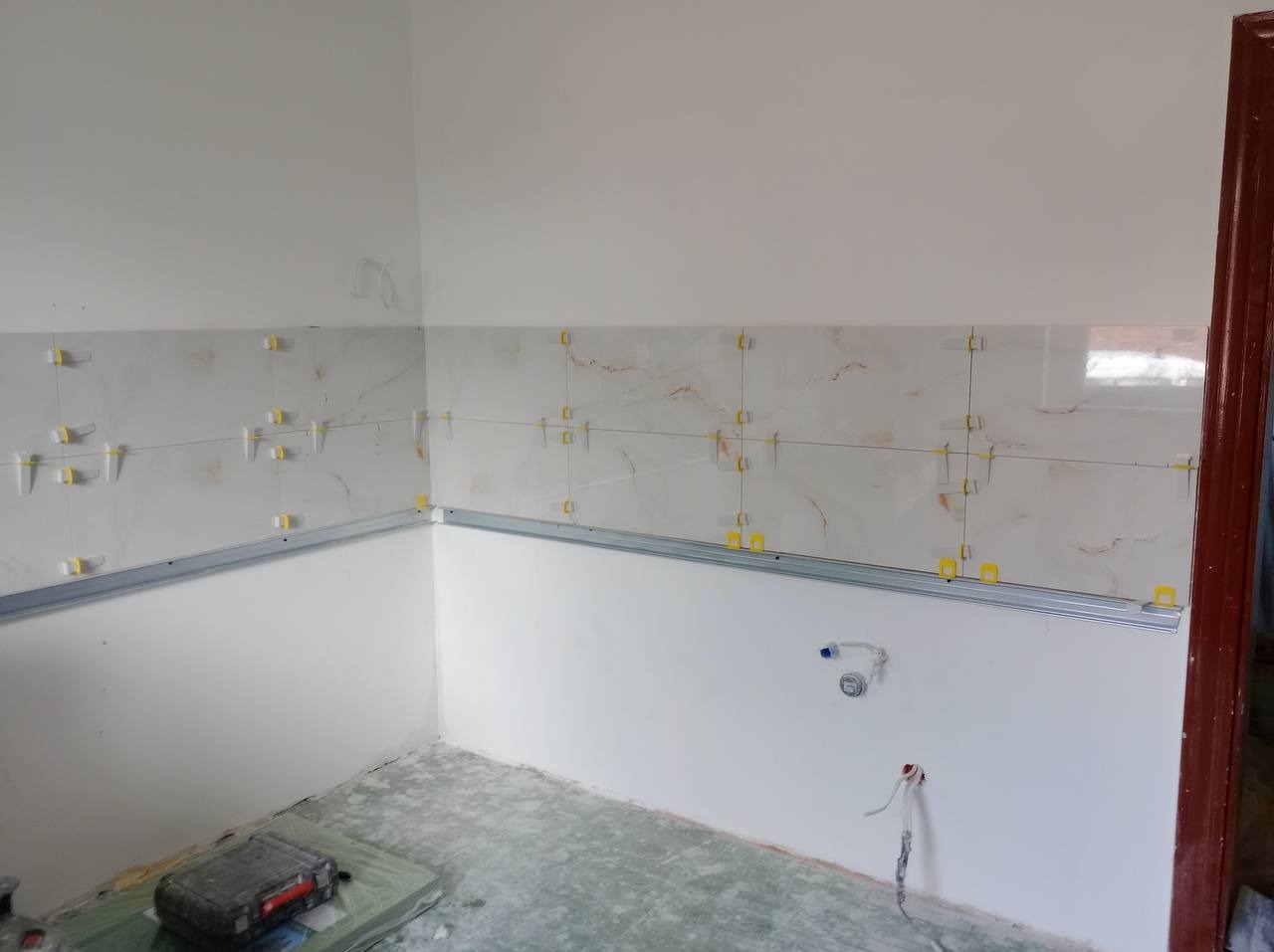 Ściana w trakcie układania płytek z użyciem listwy montażowej i krzyżyków dystansowych. Widoczne wystające kable i rury instalacyjne.