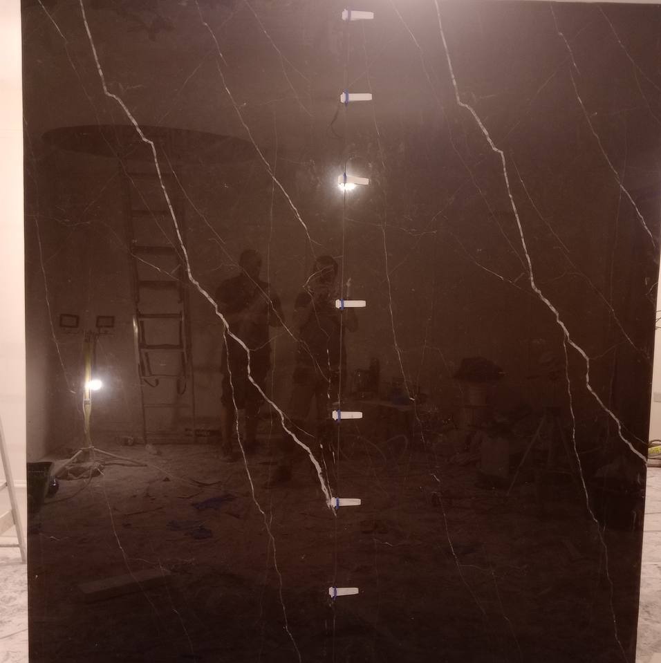 Duża płyta z ciemnego marmuru z białymi żyłami, ustawiona pionowo, z widocznymi elementami mocującymi i odbiciem sylwetek osób w tle.