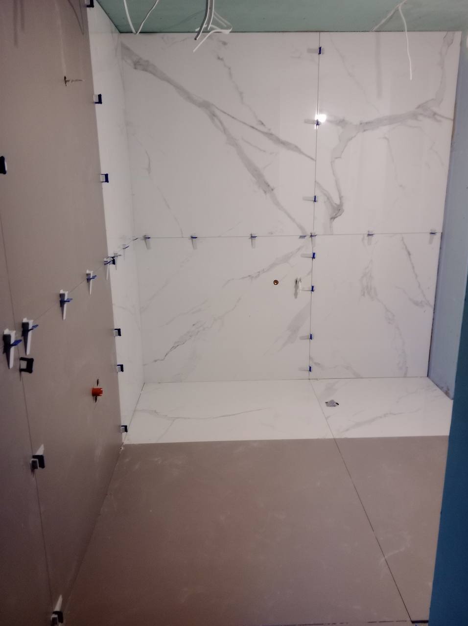 Wnętrze łazienki w trakcie remontu: białe płytki imitujące marmur na ścianach, z niebieskimi krzyżykami dystansowymi, szare ściany boczne i podłoga w trakcie prac.