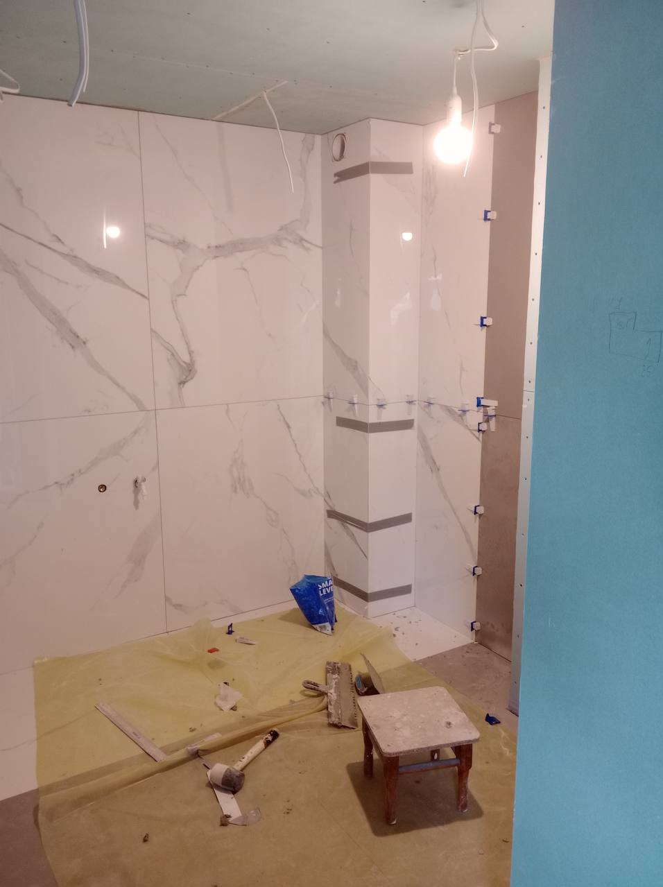 Wnętrze łazienki w trakcie wykańczania, widoczne białe płytki imitujące marmur na ścianach, narożnik obłożony płytkami z szarymi akcentami, narzędzia glazurnicze i poziomica na podłodze...