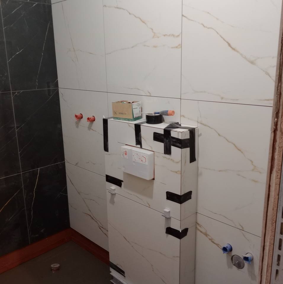 Ściana łazienki wyłożona białymi płytkami imitującymi marmur, z widoczną zabudową podtynkową WC zabezpieczoną czarną taśmą, oraz wystającymi elementami instalacji wodnej.