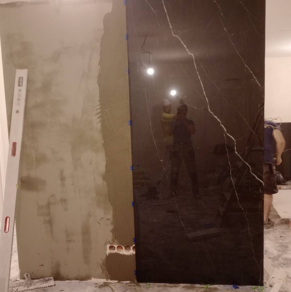 Montaż dużej, czarnej płyty imitującej marmur na ścianie, widoczna warstwa kleju i poziomica, odbicie osób w polerowanej powierzchni.