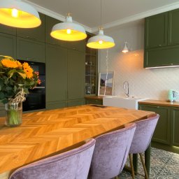 Malko Projekty Wnętrz - Kuchnia w odcieniach zieleni z drewnianym blatem stołu ułożonym we wzór jodełki, bukiet słoneczników w wazonie, biała armatura i pastelowe krzesła z weluru