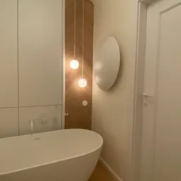 Nowoczesna łazienka z wolnostojącą białą wanną, drewnianymi akcentami i okrągłymi lampami wiszącymi. Minimalistyczny design i jasna kolorystyka.