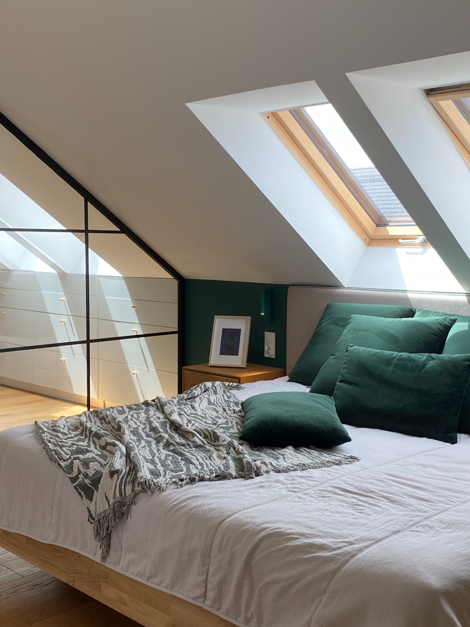 Sypialnia na poddaszu z łóżkiem z drewnianą ramą, zielonymi poduszkami i pledem w biało-szare wzory. W tle biała zabudowa szaf z czarnymi ramami i okna dachowe wpuszczające światło.