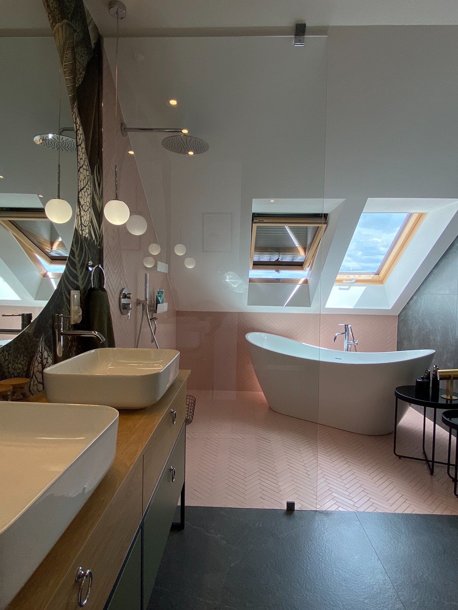 Jasna łazienka na poddaszu z dwoma umywalkami nablatowymi na drewnianej szafce, wolnostojącą wanną pod oknami dachowymi i różową mozaiką na ścianach oraz podłodze.