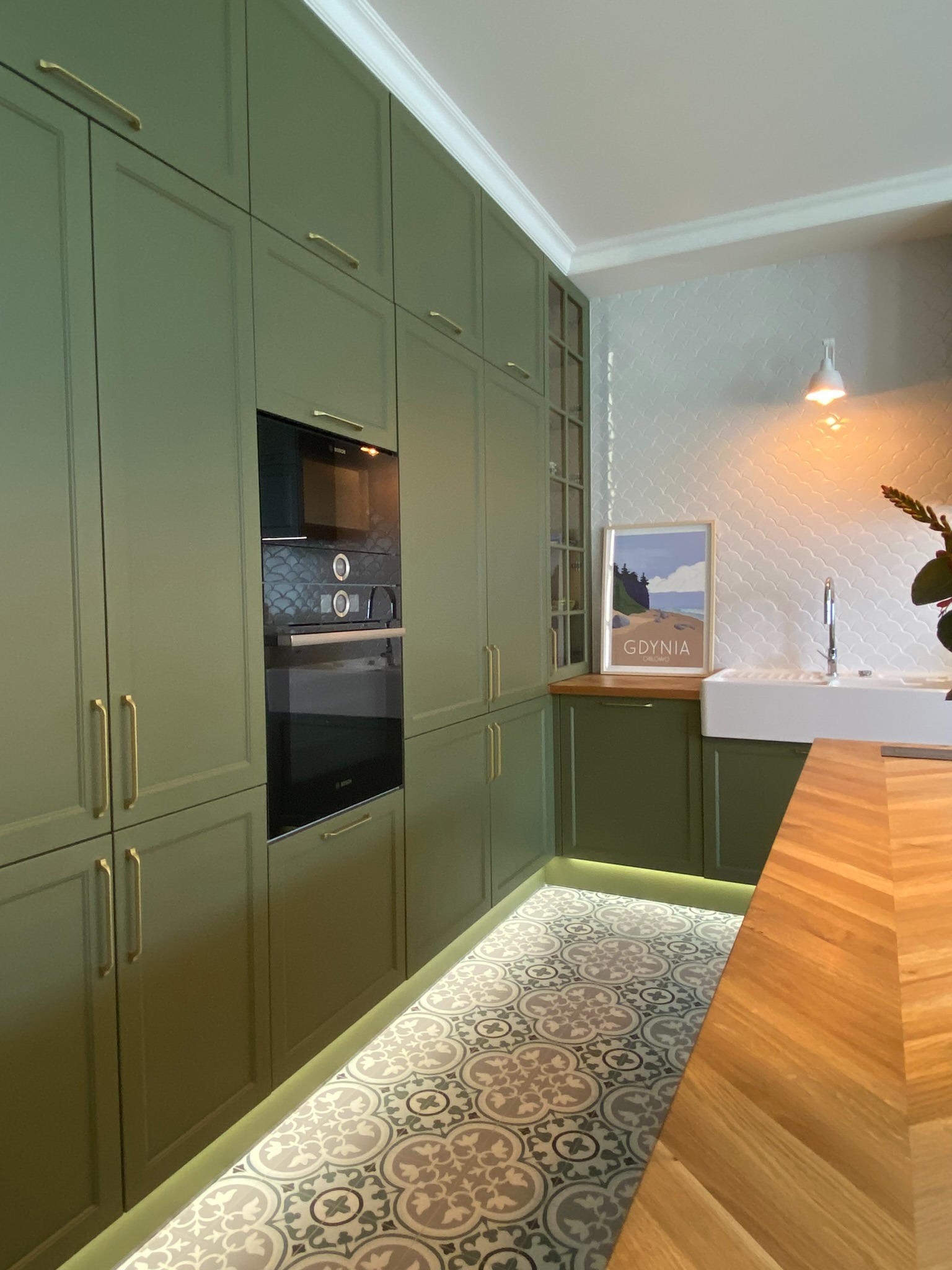 Kuchnia w odcieniach zieleni z drewnianym blatem, wzorzystą podłogą i oświetleniem LED pod szafkami, plakat z motywem Gdyni na blacie.