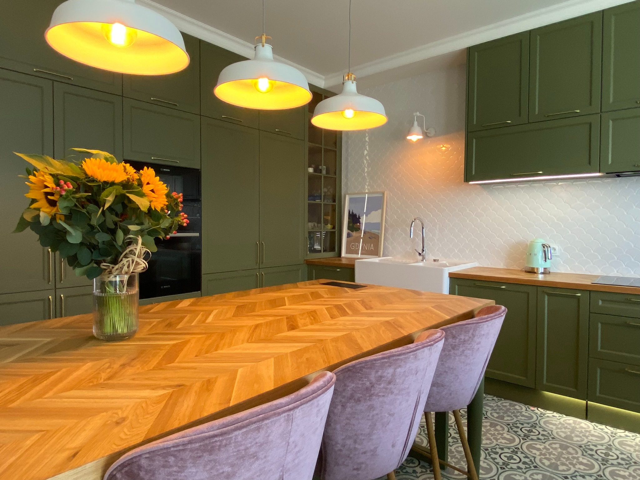 Kuchnia w odcieniach zieleni z drewnianym blatem stołu ułożonym we wzór jodełki, bukiet słoneczników w wazonie, biała armatura i pastelowe krzesła z weluru