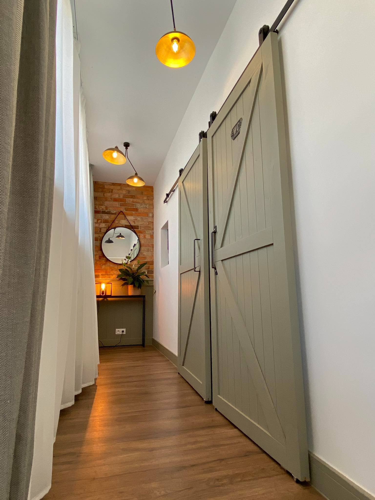 Korytarz z drewnianą podłogą, przesuwne drzwi w stylu stodoły, ceglana ściana z okrągłym lustrem i konsolą z rośliną, oświetlenie sufitowe z ciepłym światłem.
