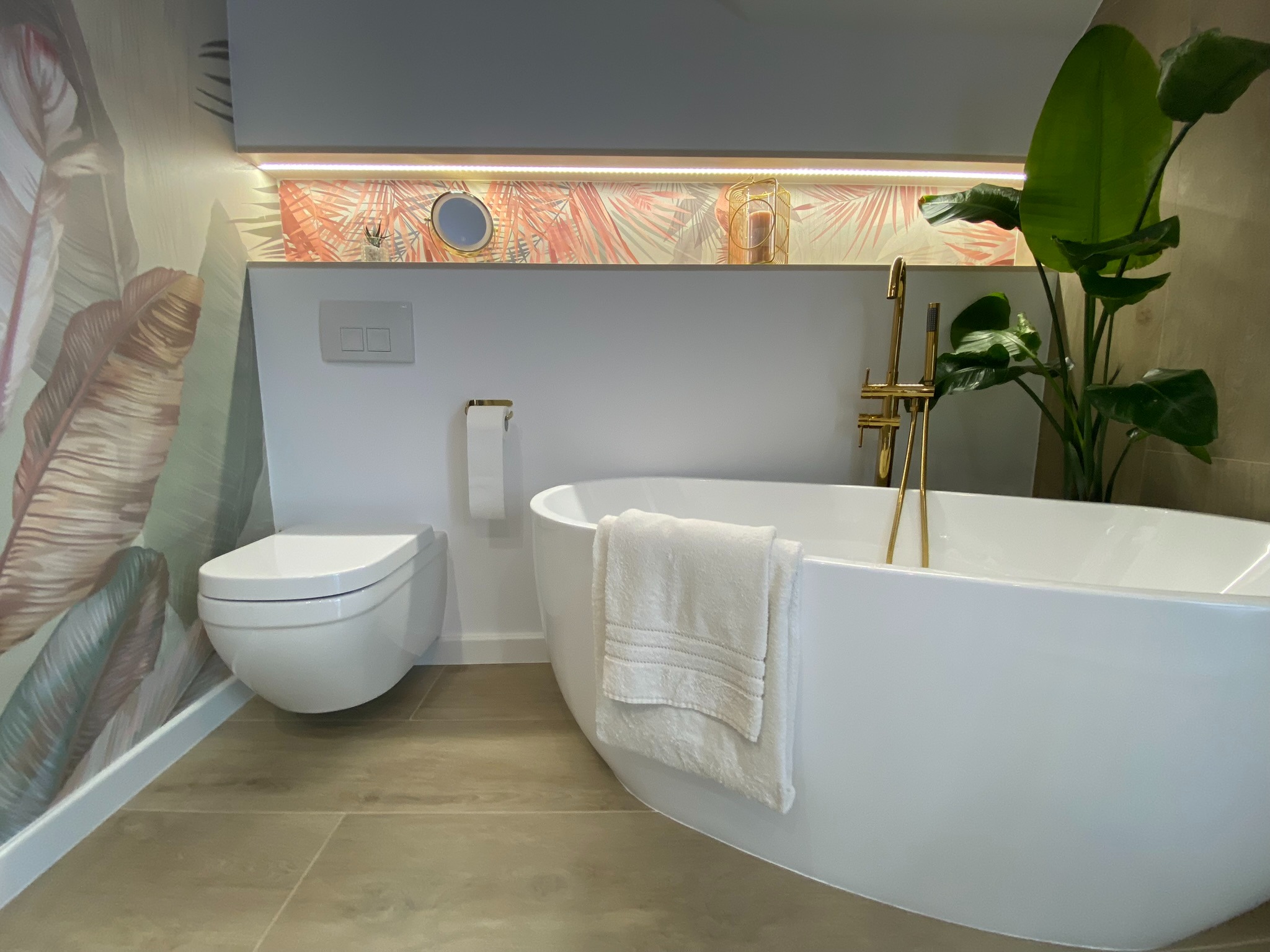 Nowoczesna łazienka z wolnostojącą białą wanną, złotą armaturą, wiszącą toaletą i tapetą w motywy roślinne, oświetlona listwą LED.