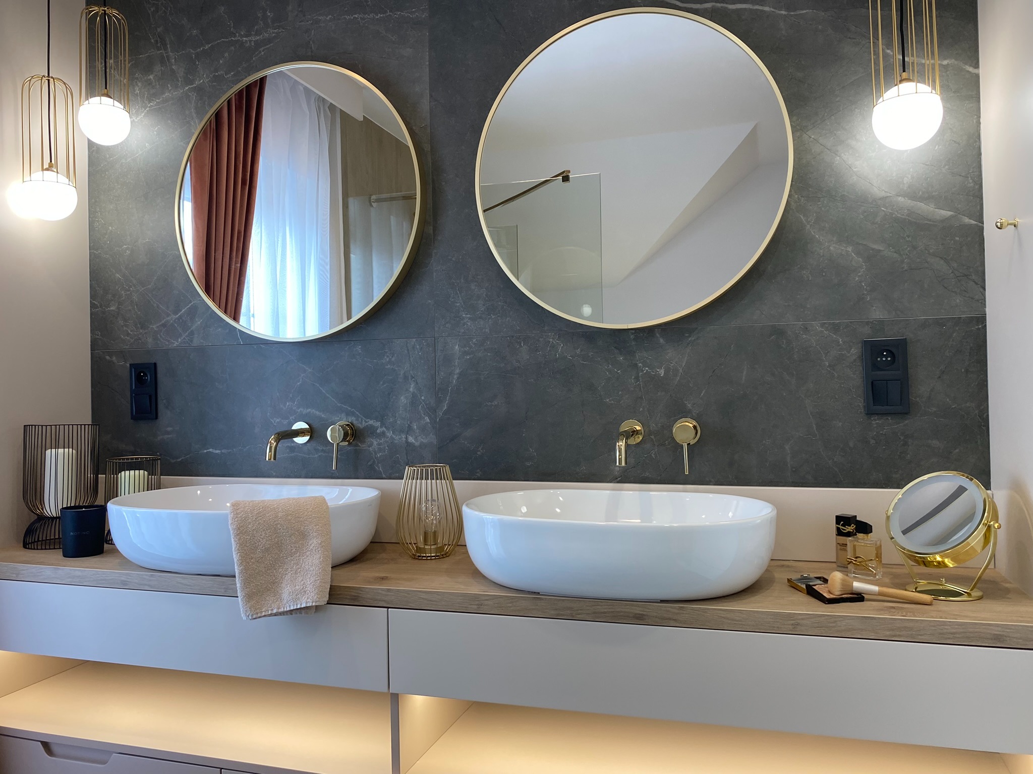 Nowoczesna łazienka z dwoma okrągłymi lustrami w złotych ramach, wiszącymi lampami z metalowymi kloszami, marmurowymi płytkami ściennymi oraz dwiema białymi umywalkami nablatowymi na drewnianym...
