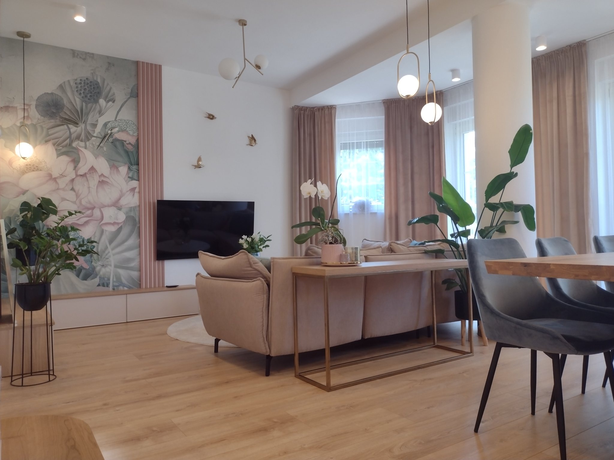 Jasny salon z fototapetą w kwiaty, beżową sofą, drewnianą podłogą i szarymi krzesłami przy stole z drewnianym blatem, oświetlony lampami wiszącymi.