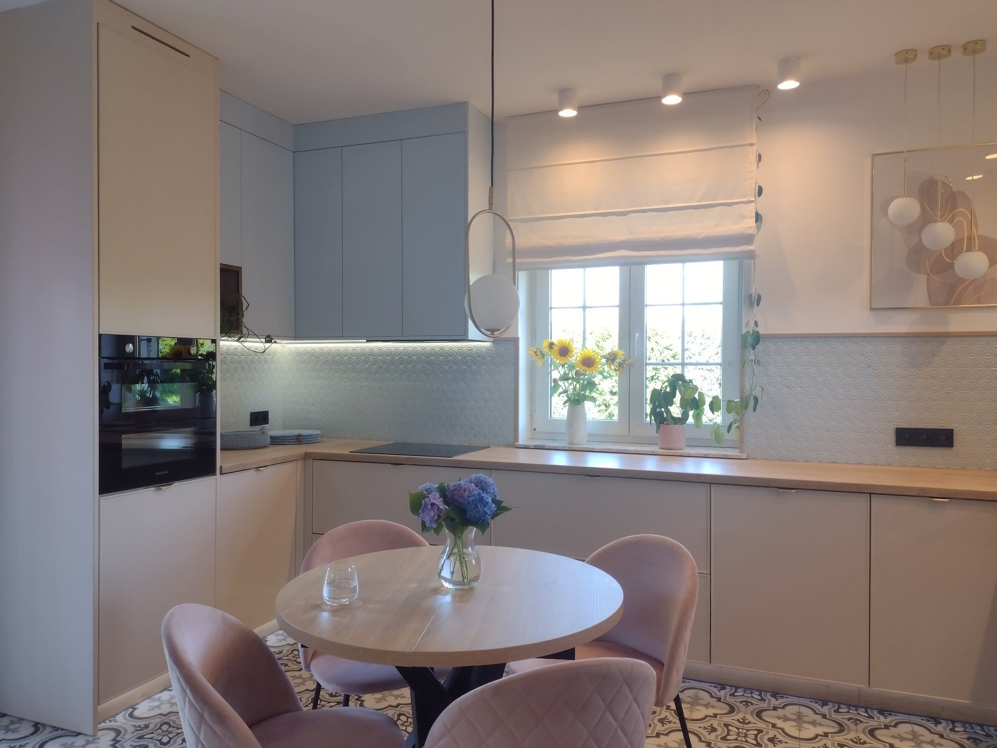 Jasna kuchnia z okrągłym stołem i różowymi krzesłami, kwiaty w wazonie, wzorzysta podłoga, minimalistyczne oświetlenie.