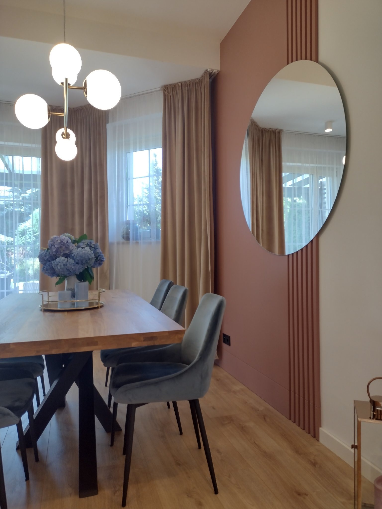Jadalnia w stylu nowoczesnym z drewnianym stołem, szarymi krzesłami, bukietem hortensji, okrągłym lustrem i lampą sufitową z kulistymi kloszami.