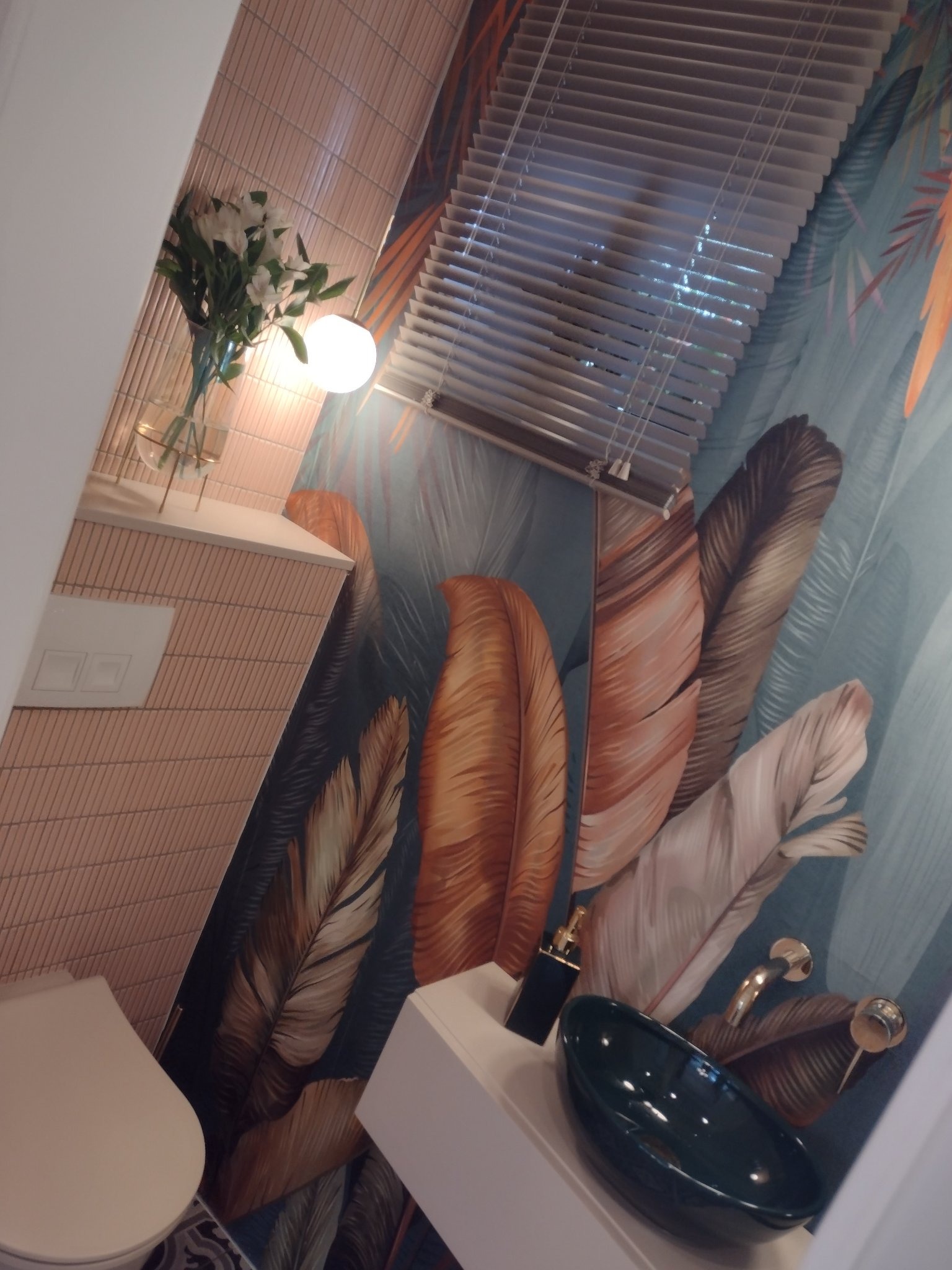 Nowoczesna łazienka z umywalką w kolorze butelkowej zieleni, armaturą w odcieniu złota, tapetą w tropikalne liście i roletami w oknie.