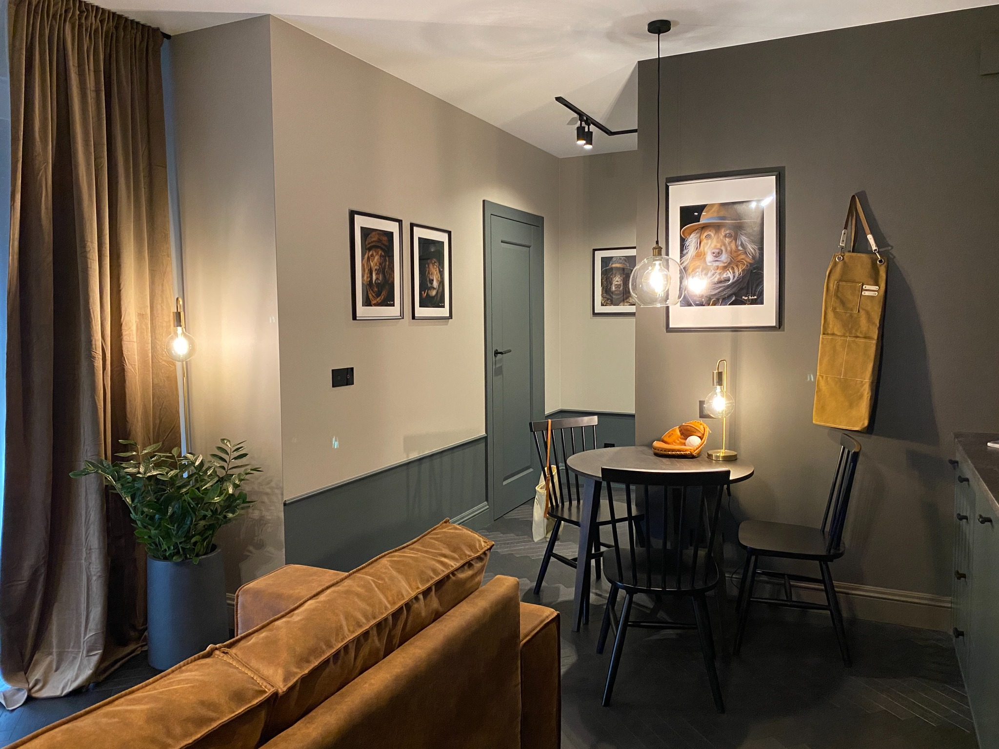 Nowoczesne wnętrze z ciemnymi ścianami, drewnianą podłogą w jodełkę i okrągłym stołem z czarnymi krzesłami. Na ścianach wiszą obrazy psów w kapeluszach, a nad stołem wisi szklana lampa. Na stole...