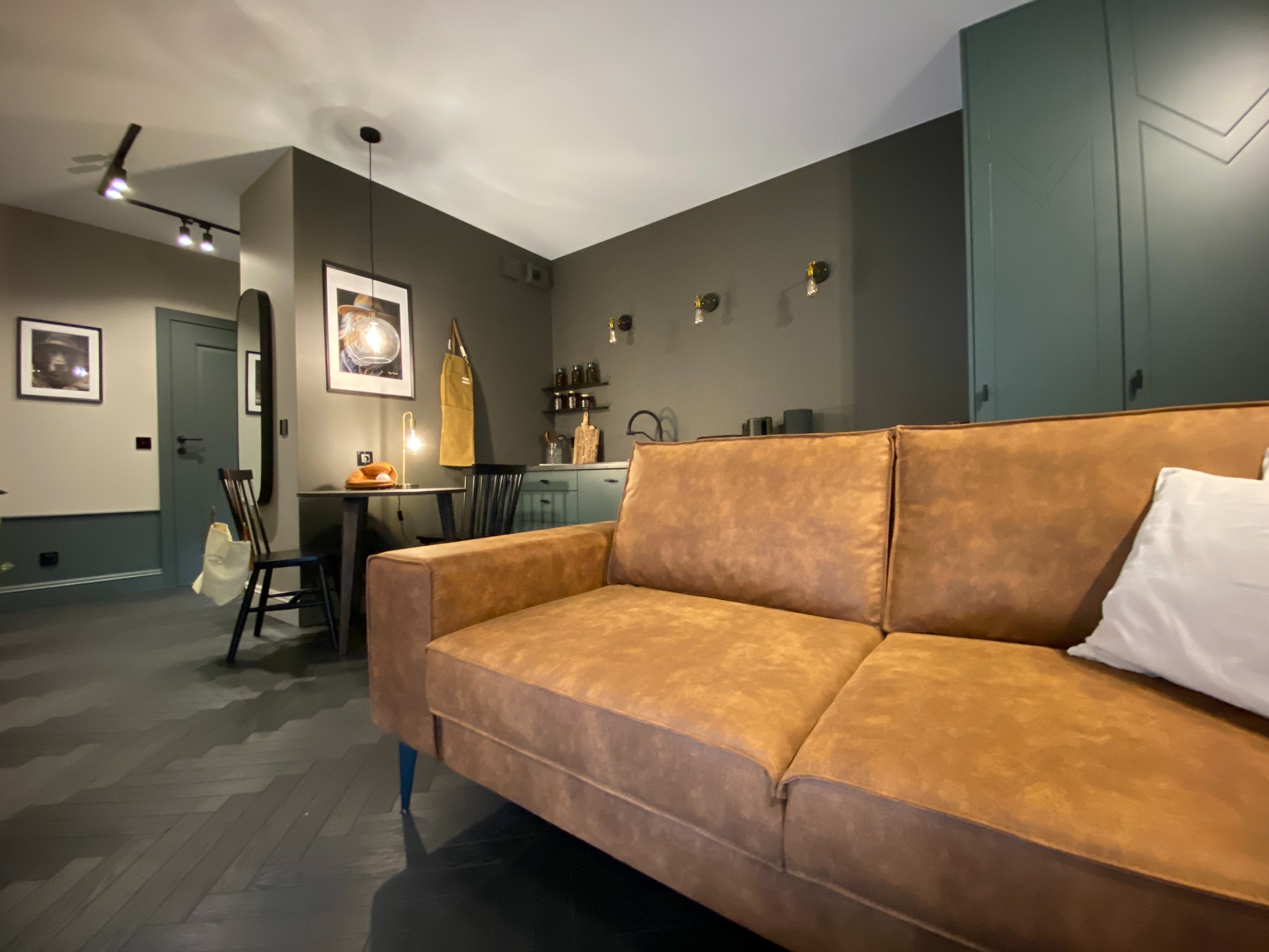 Nowoczesne wnętrze mieszkania z brązową sofą na pierwszym planie, ciemna podłoga w jodełkę, w tle aneks kuchenny i stół z krzesłami, oświetlenie punktowe i wisząca lampa.