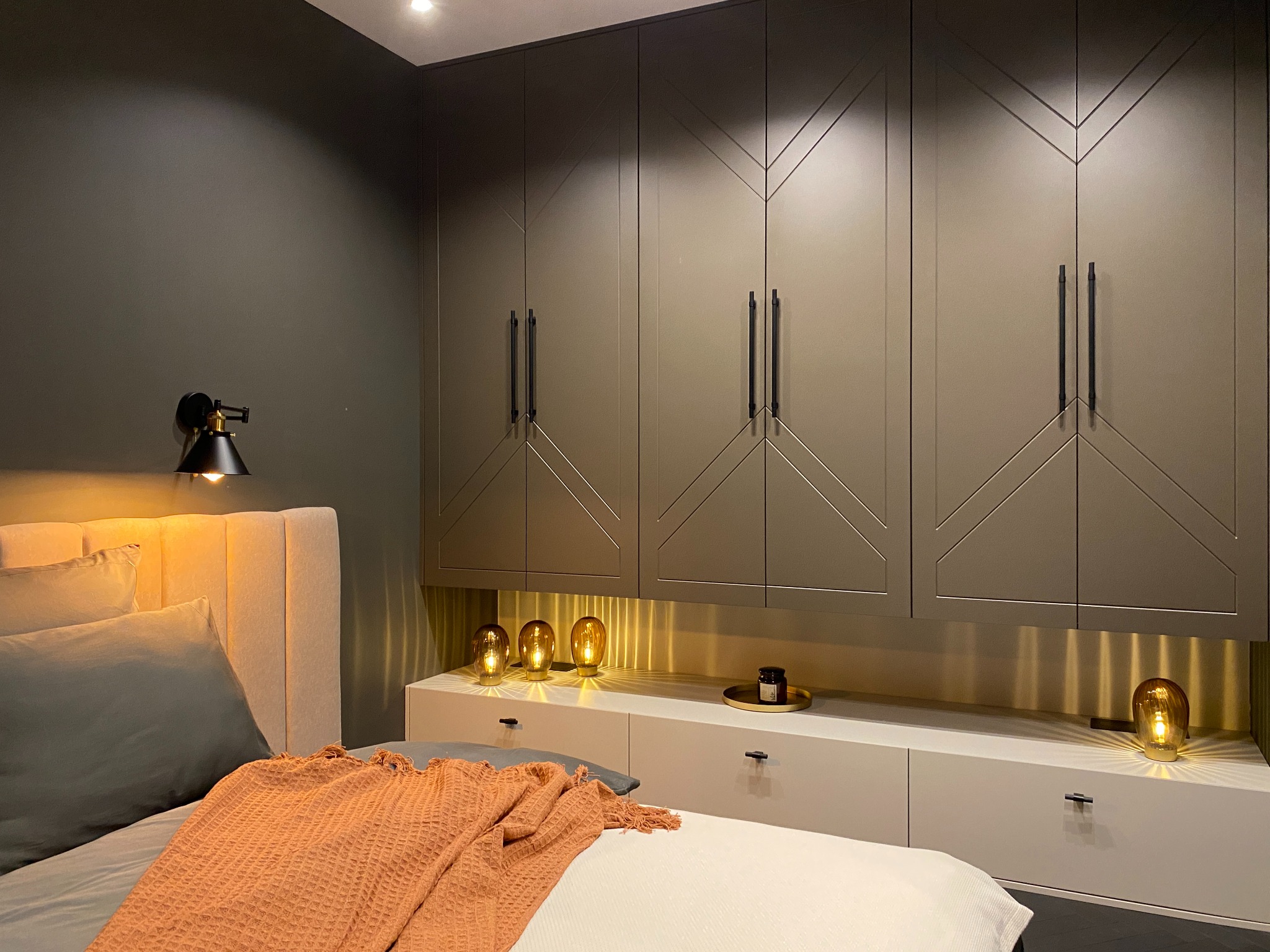 Nowoczesna sypialnia z dużym łóżkiem, szarą pościelą i pomarańczowym pledem. Nad łóżkiem zabudowa szaf z geometrycznym wzorem. Na szafce nocnej dekoracyjne lampki w kształcie żarówek.