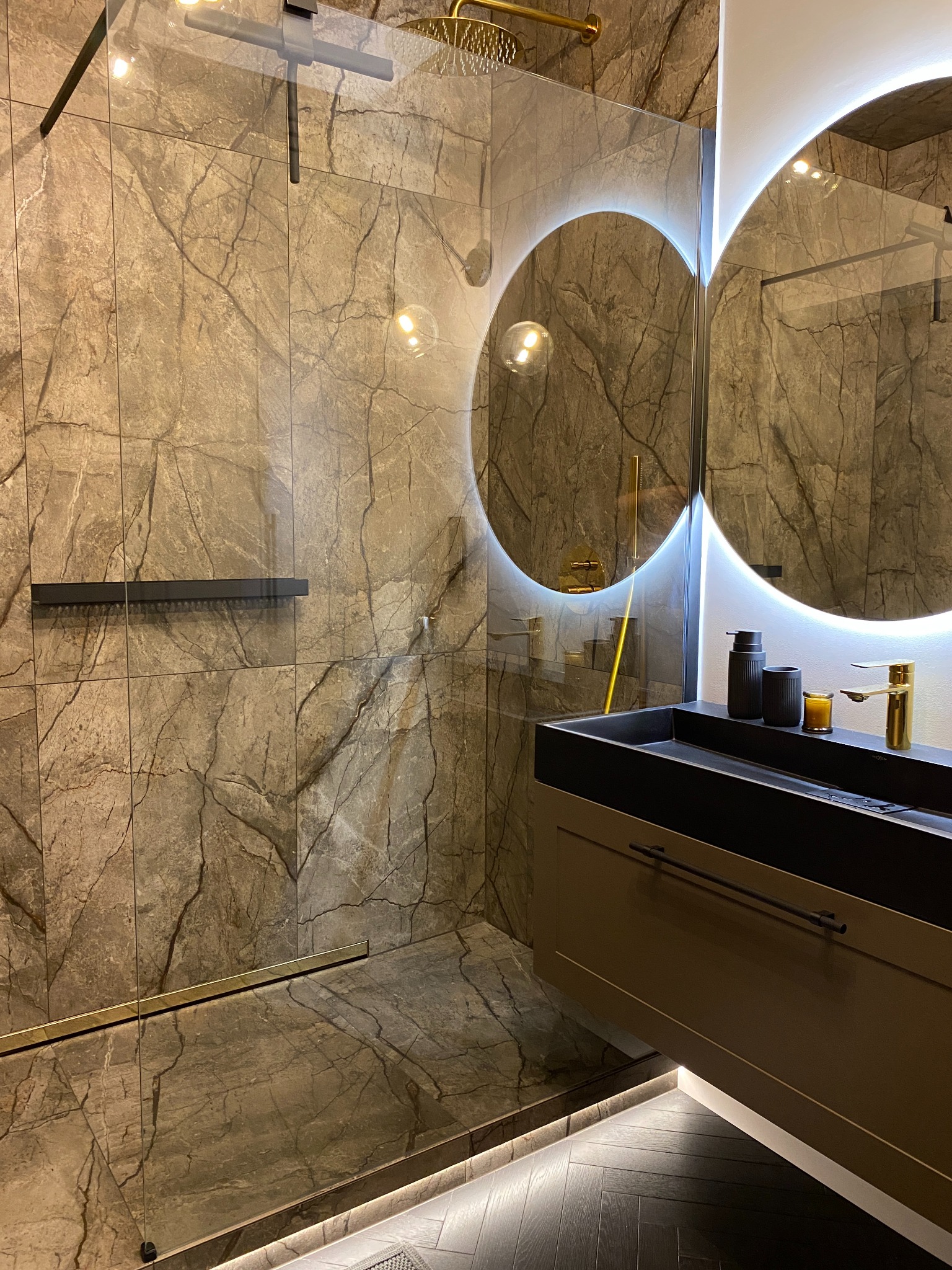 Nowoczesna łazienka z kabiną prysznicową typu walk-in, wykończona płytkami imitującymi marmur z wyraźnym żyłowaniem, podświetlanymi okrągłymi lustrami nad czarną umywalką i złotą armaturą,...