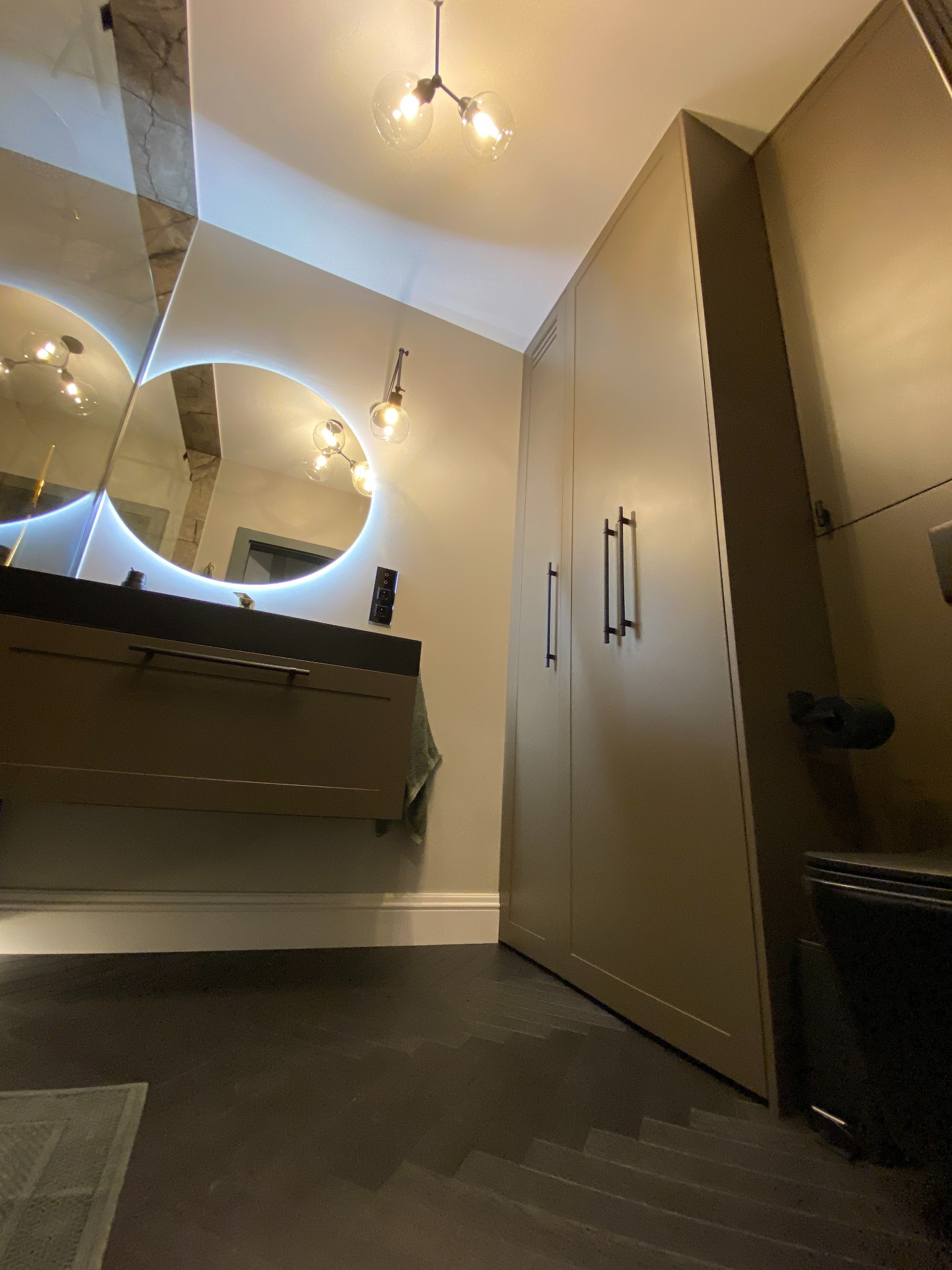 Nowoczesna łazienka z okrągłym lustrem podświetlanym LED, szafką pod umywalką w kolorze taupe, wysoką szafą i parkietem w jodełkę.