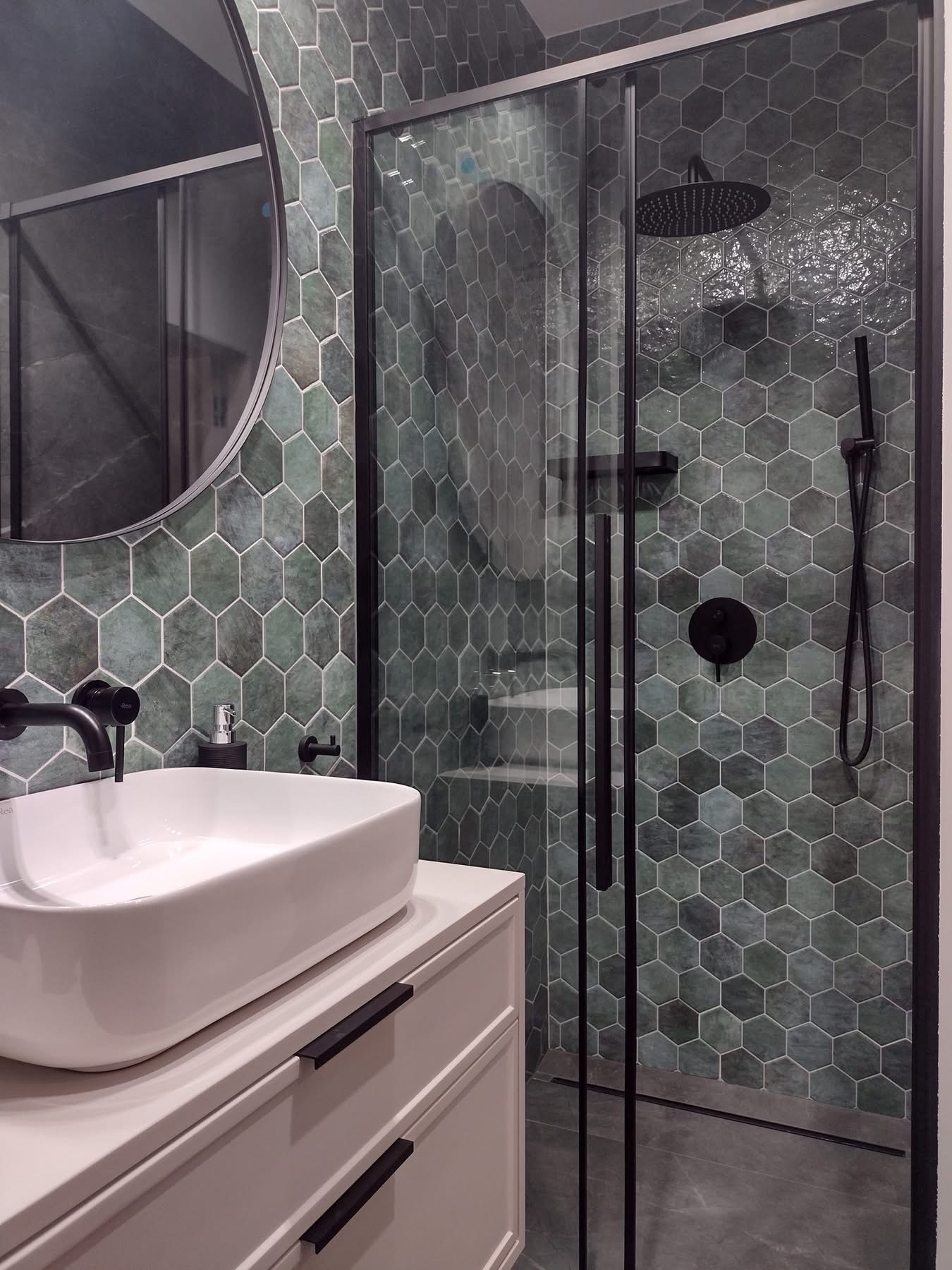 Nowoczesna łazienka z heksagonalnymi, zielonymi płytkami, czarną armaturą i kabiną prysznicową. Biała umywalka nablatowa na szafce. Okrągłe lustro.
