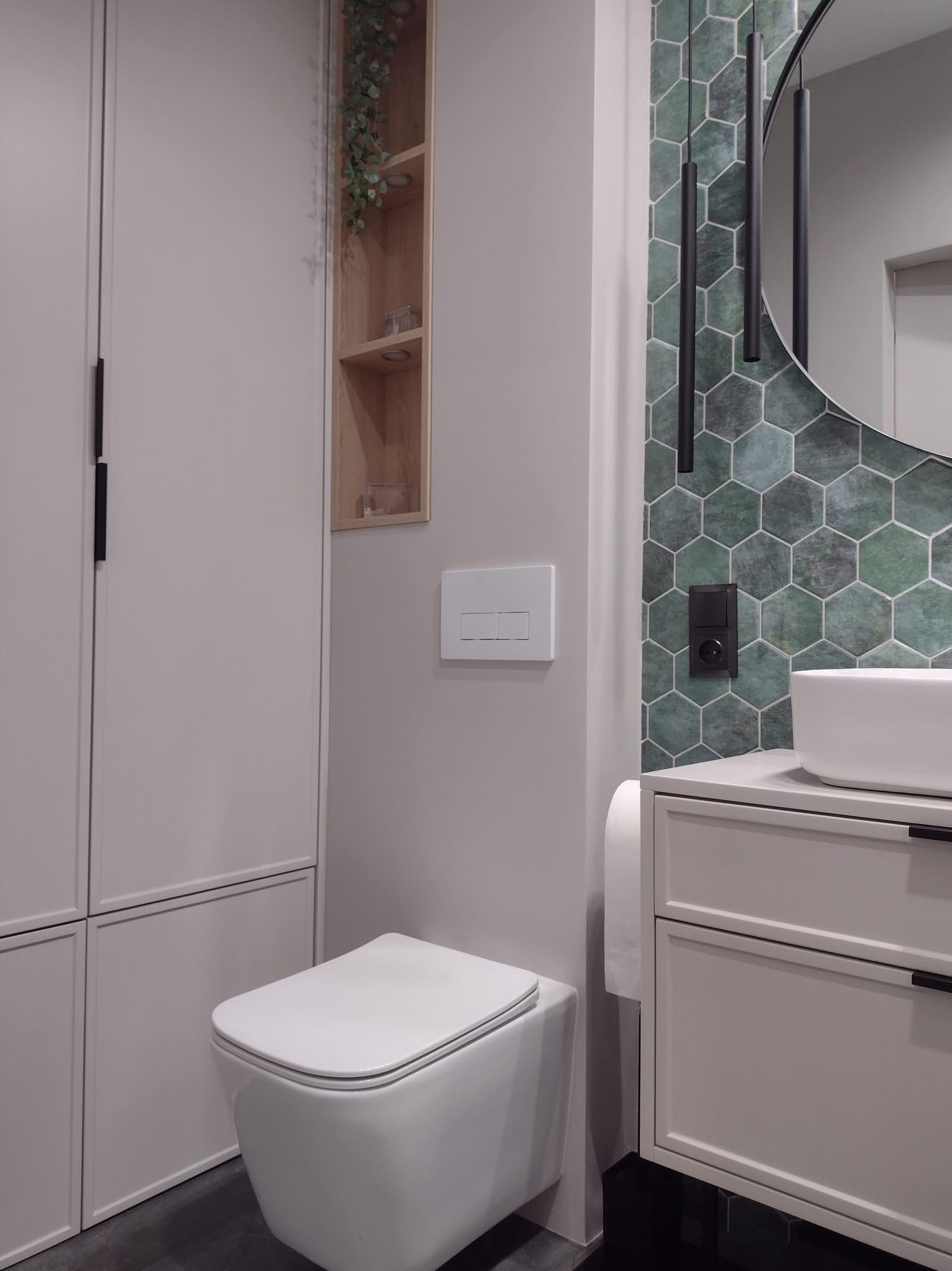 Nowoczesna aranżacja łazienki w Kątach Wrocławskich: wisząca toaleta, szafka z umywalką i lustro na tle ściany z heksagonalnymi płytkami w odcieniach zieleni.