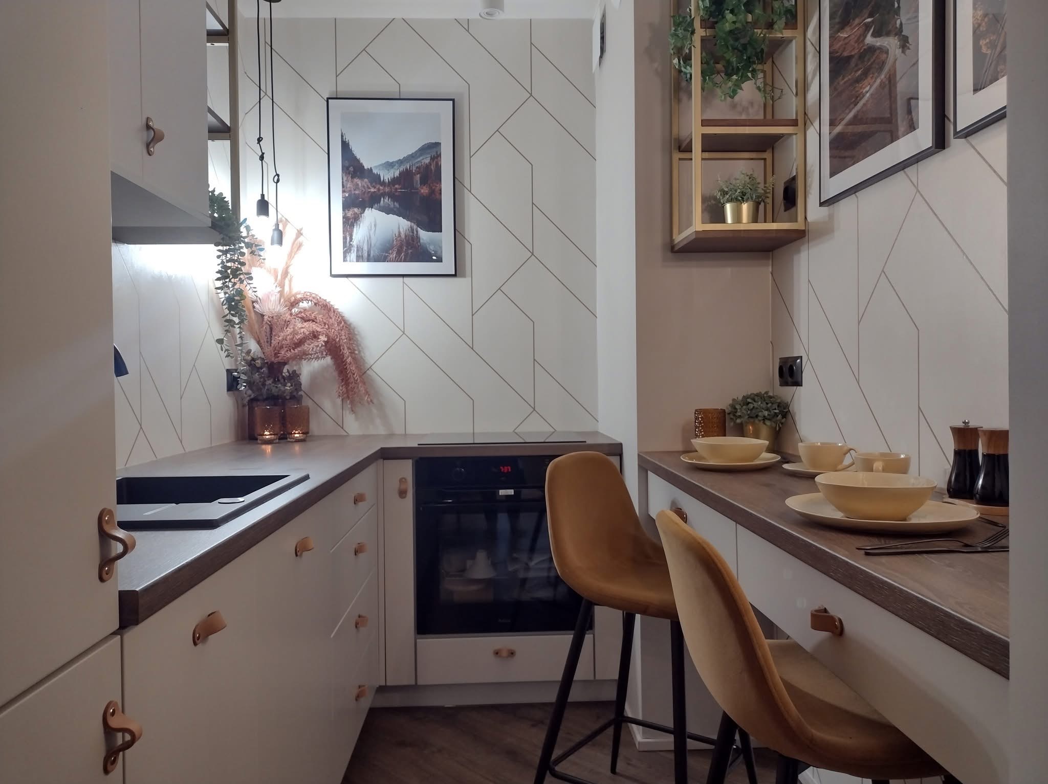 Jasna kuchnia z drewnianym blatem, ozdobnymi uchwytami i geometryczną tapetą w romby. Dwa krzesła barowe i dekoracje roślinne dopełniają aranżację.