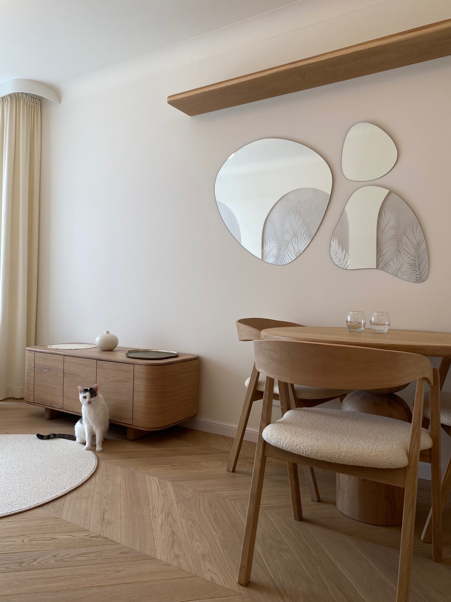 Jasne wnętrze jadalni z drewnianym stołem, krzesłami i komodą. Na ścianie asymetryczne lustra. Uwagę przykuwa siedzący kot o umaszczeniu biało-czarnym.