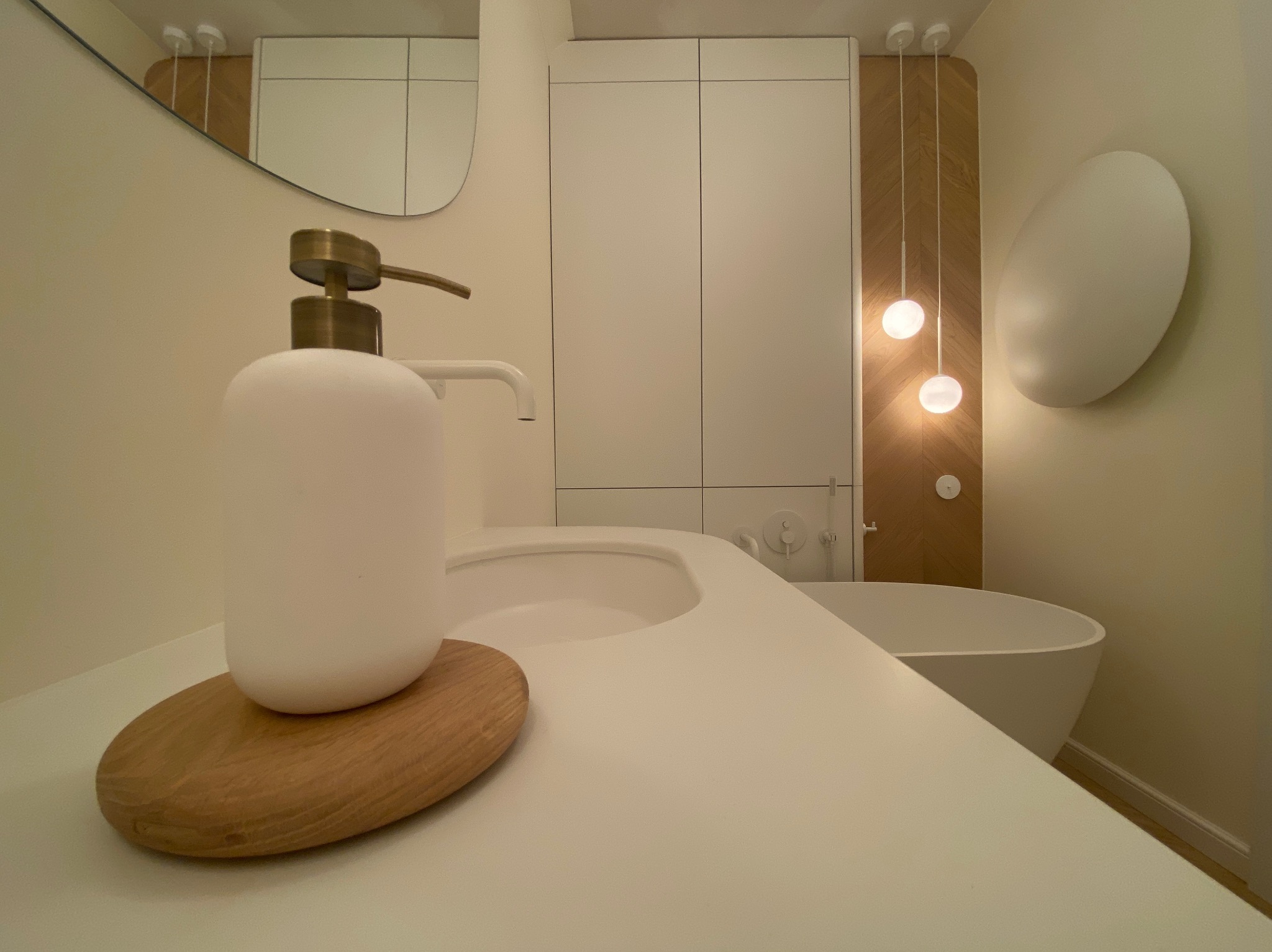 Minimalistyczna łazienka: biała umywalka z dozownikiem na drewnianej podstawce, owalne lustro i lampy wiszące na tle drewnianej ściany. Nowoczesny design.