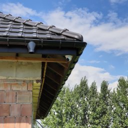 ELFOT GRZEGORZ PALIŃSKI - Kamera kopułkowa zamontowana pod okapem dachu, widok z dołu na tle błękitnego nieba i zielonych drzew, widoczne elementy konstrukcyjne budynku w trakcie budowy.