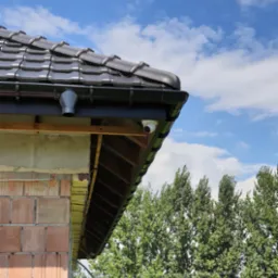 Kamera kopułkowa zamontowana pod okapem dachu, widok z dołu na tle błękitnego nieba i zielonych drzew, widoczne elementy konstrukcyjne budynku w trakcie budowy.