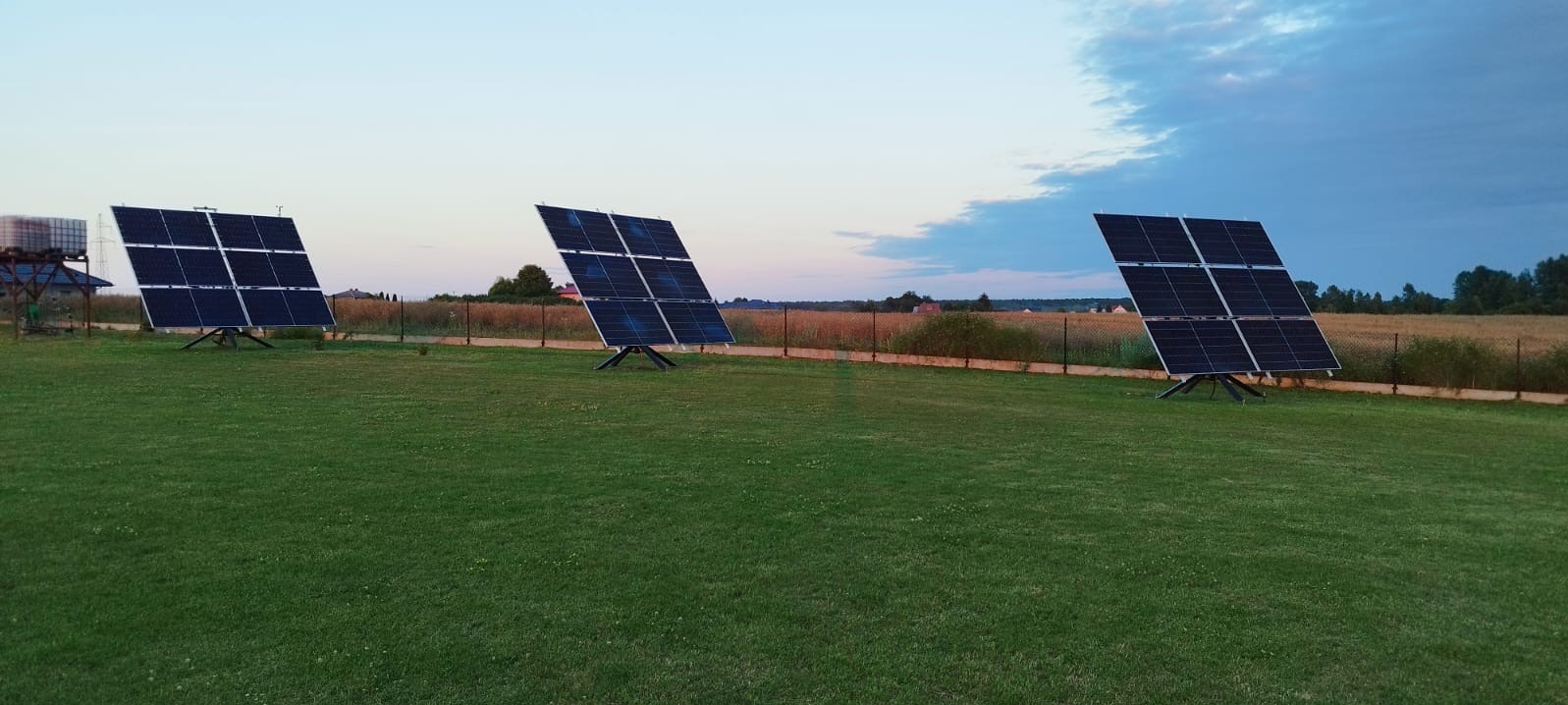 Trzy panele fotowoltaiczne ustawione na trawniku, widoczne na tle pola i nieba o zmierzchu.